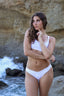 Woman in a white bikini standing on a rocky beach with ocean waves.